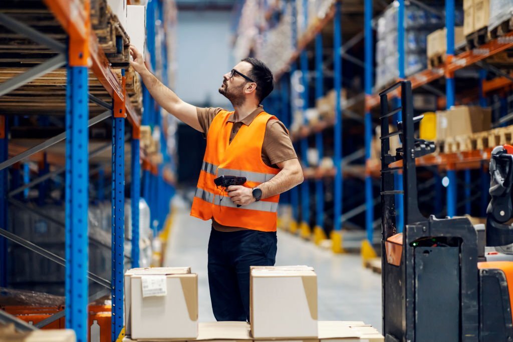 Man inside warehouse, reaching for a box above head on a shelve 