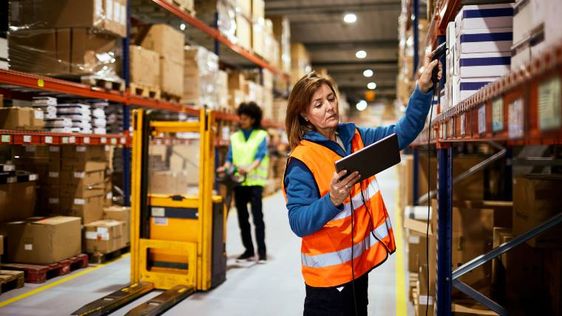 Woman in a safety vest reaching for a box in a warehouse.